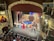 A vibrant theater stage with a group of performers dressed in costumes, holding a large flag, possibly from a staged performance. An orchestra is visible in the pit below the stage. The theater's interior is ornate, with elaborate decorations and balconies.