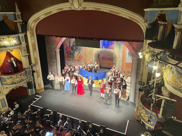 A vibrant theater stage with a group of performers dressed in costumes, holding a large flag, possibly from a staged performance. An orchestra is visible in the pit below the stage. The theater's interior is ornate, with elaborate decorations and balconies.
