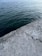 A concrete surface meets a body of water with gently rippling waves. The concrete is textured and weathered, showing signs of age and wear. The water is a deep blue-green color, suggesting depth and movement beneath the surface.