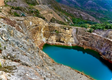 A large, turquoise lake sits nestled within rocky, mountainous terrain. The surrounding landscape is rugged, with patches of greenery and barren, uneven rock formations. The area appears remote and untouched, giving a sense of natural beauty and isolation.