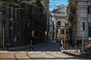 A bustling urban street with historic architecture flanking each side. The streetcar tracks run through the middle, while pedestrians cross and vehicles drive in the background. The scene is set under a clear sky with a prominent domed building visible through an archway in the distance.