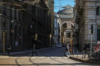 A bustling urban street with historic architecture flanking each side. The streetcar tracks run through the middle, while pedestrians cross and vehicles drive in the background. The scene is set under a clear sky with a prominent domed building visible through an archway in the distance.