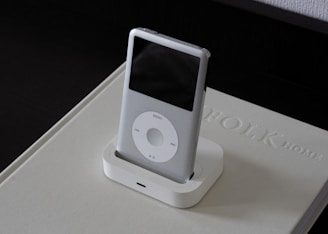 A classic MP3 player with a sleek white design is placed in a docking station on top of a beige book with embossed text. The device features a scroll wheel and a small screen, and it is set against a dark background.