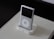 A classic MP3 player with a sleek white design is placed in a docking station on top of a beige book with embossed text. The device features a scroll wheel and a small screen, and it is set against a dark background.