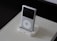 A classic MP3 player with a sleek white design is placed in a docking station on top of a beige book with embossed text. The device features a scroll wheel and a small screen, and it is set against a dark background.