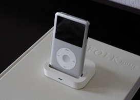 A classic MP3 player with a sleek white design is placed in a docking station on top of a beige book with embossed text. The device features a scroll wheel and a small screen, and it is set against a dark background.