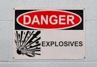 A warning sign with a bold red and white design that prominently displays the word 'DANGER' in an oval shape. Below, the sign reads 'EXPLOSIVES' in bold black letters against a white background. On the left side, there is a graphic depicting an explosion with black stylized fragments emanating from a central point. The sign is mounted on a light-colored brick wall.