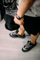 A person wearing casual sneakers stands on a light-colored wooden floor, holding a black cap in their hand. In the background, there are clothes hanging, showing prints with tropical designs.
