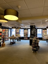 A spacious, well-lit bookstore with large windows allowing natural light to illuminate the space. Shelves are lined with a variety of books, and tables display stacks of books for browsing. Overhead, large circular ceiling lights contribute to the warm ambiance.