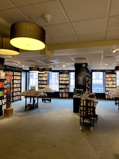 A spacious, well-lit bookstore with large windows allowing natural light to illuminate the space. Shelves are lined with a variety of books, and tables display stacks of books for browsing. Overhead, large circular ceiling lights contribute to the warm ambiance.