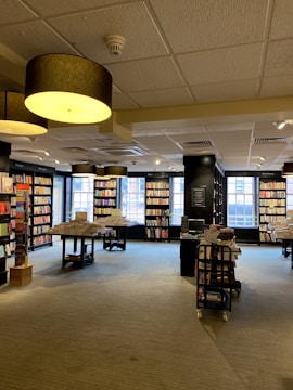 A spacious, well-lit bookstore with large windows allowing natural light to illuminate the space. Shelves are lined with a variety of books, and tables display stacks of books for browsing. Overhead, large circular ceiling lights contribute to the warm ambiance.