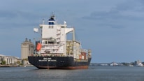 A large cargo ship sails on a calm body of water with numerous shipping containers stacked on its deck. In the background, there is a clear view of a cityscape featuring modern buildings, including a prominent structure labeled 'Savannah Courthouse' and a hotel. The sky is slightly overcast with scattered clouds.