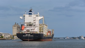 A large cargo ship sails on a calm body of water with numerous shipping containers stacked on its deck. In the background, there is a clear view of a cityscape featuring modern buildings, including a prominent structure labeled 'Savannah Courthouse' and a hotel. The sky is slightly overcast with scattered clouds.