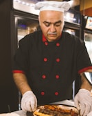 A chef in a black uniform with red accents and a white hat is carefully arranging food on a plate. The chef is wearing white gloves, focusing intently on the task. The background features kitchen appliances and shelving, suggesting a professional kitchen setting.