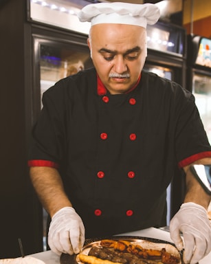 A chef in a black uniform with red accents and a white hat is carefully arranging food on a plate. The chef is wearing white gloves, focusing intently on the task. The background features kitchen appliances and shelving, suggesting a professional kitchen setting.