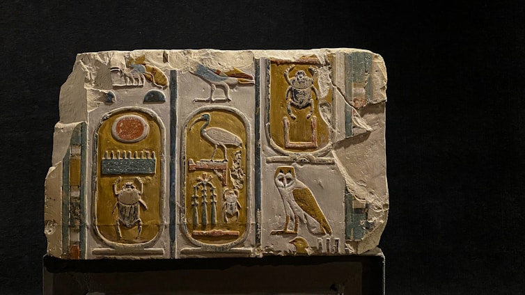 A stone tablet featuring ancient Egyptian hieroglyphics with various symbols including birds, a scarab, an owl, and other symbolic figures. The carvings are colored with earthy tones such as yellow, and brown against a light stone background. The tablet appears aged and slightly damaged, adding to its historical charm.