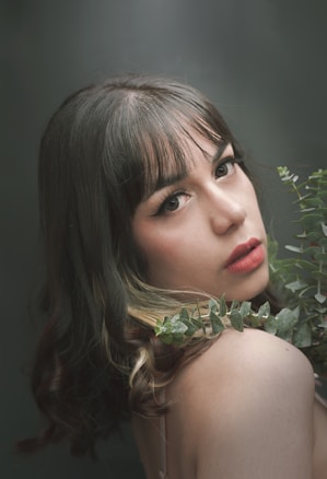 A woman with long, wavy hair and bangs looks over her shoulder, surrounded by green foliage. Her makeup is soft and natural, with a focus on her eyes and lips. The background is a soft gradient of greens, complementing the plants.