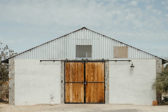 A rustic building with a large wooden sliding barn door in the center, flanked by two stone pillars. The upper half of the building is constructed from corrugated metal, featuring two small windows. A potted plant and a light fixture are visible on the exterior.