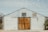 A rustic building with a large wooden sliding barn door in the center, flanked by two stone pillars. The upper half of the building is constructed from corrugated metal, featuring two small windows. A potted plant and a light fixture are visible on the exterior.
