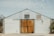 A rustic building with a large wooden sliding barn door in the center, flanked by two stone pillars. The upper half of the building is constructed from corrugated metal, featuring two small windows. A potted plant and a light fixture are visible on the exterior.