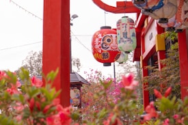 A vibrant scene featuring traditional Japanese lanterns hanging from a red wooden structure. The lanterns display colorful designs with one prominently featuring a stylized face. Surrounding the structure are lush flowers and plants, contributing to an atmosphere reminiscent of a serene garden. The background includes additional decorative elements and foliage.
