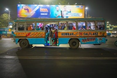 A crowded blue and yellow bus with a mix of passengers inside is driving through a city street at night. The bus has large signages for various destinations including Howrah and Saltlake. Above the bus, there is a brightly lit billboard featuring people in colorful outfits.