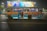 A crowded blue and yellow bus with a mix of passengers inside is driving through a city street at night. The bus has large signages for various destinations including Howrah and Saltlake. Above the bus, there is a brightly lit billboard featuring people in colorful outfits.
