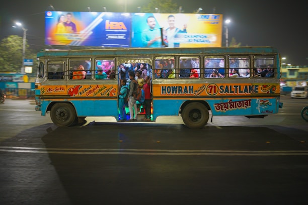 A crowded blue and yellow bus with a mix of passengers inside is driving through a city street at night. The bus has large signages for various destinations including Howrah and Saltlake. Above the bus, there is a brightly lit billboard featuring people in colorful outfits.