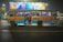 A crowded blue and yellow bus with a mix of passengers inside is driving through a city street at night. The bus has large signages for various destinations including Howrah and Saltlake. Above the bus, there is a brightly lit billboard featuring people in colorful outfits.