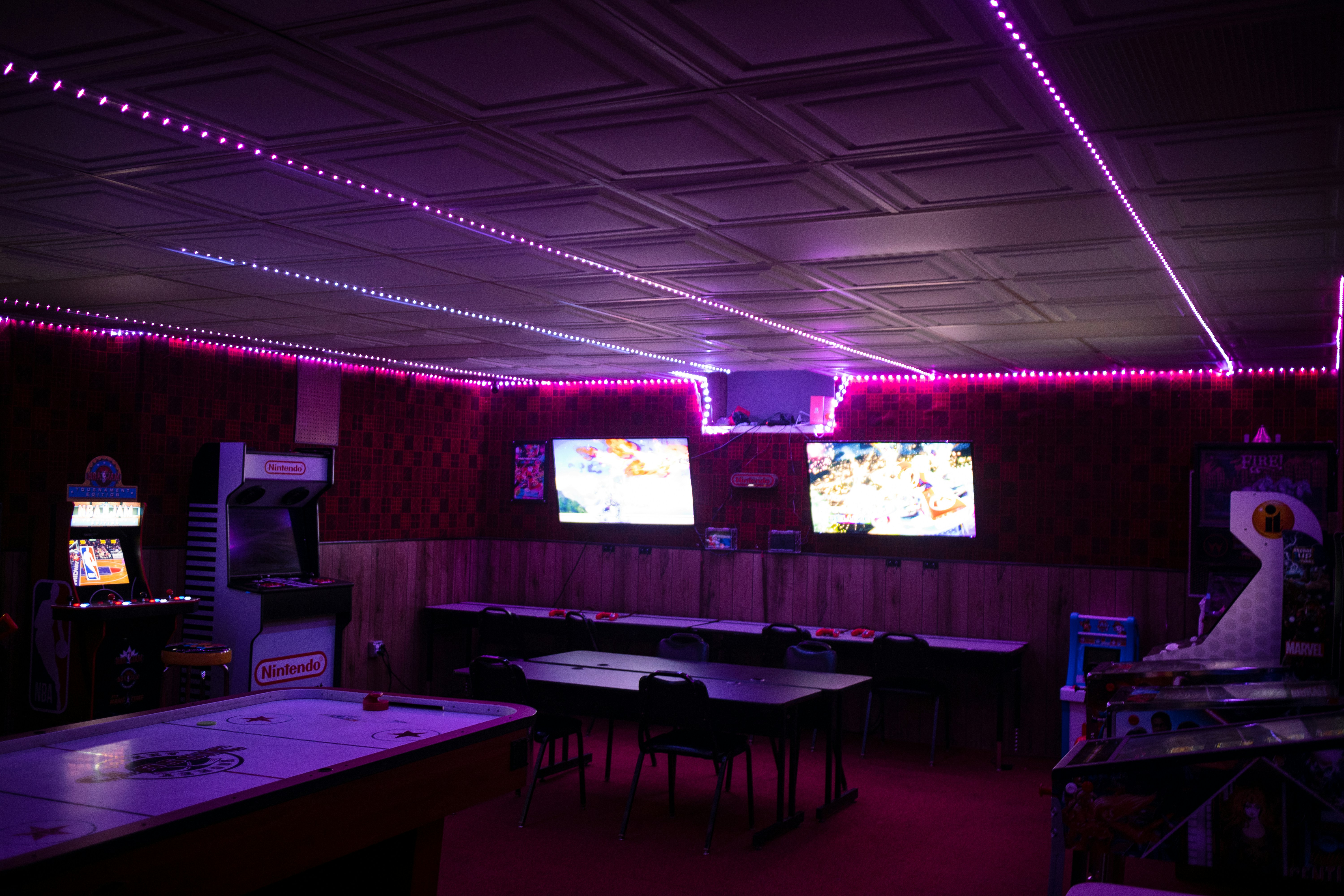 A dimly lit room with vibrant purple LED strip lights on the ceiling, featuring multiple gaming stations. There are two large screens showing video games, along with various arcade machines, including a Nintendo arcade cabinet. An air hockey table is positioned in the foreground.