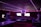 A dimly lit room with vibrant purple LED strip lights on the ceiling, featuring multiple gaming stations. There are two large screens showing video games, along with various arcade machines, including a Nintendo arcade cabinet. An air hockey table is positioned in the foreground.