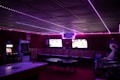 A dimly lit room with vibrant purple LED strip lights on the ceiling, featuring multiple gaming stations. There are two large screens showing video games, along with various arcade machines, including a Nintendo arcade cabinet. An air hockey table is positioned in the foreground.