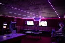 A dimly lit room with vibrant purple LED strip lights on the ceiling, featuring multiple gaming stations. There are two large screens showing video games, along with various arcade machines, including a Nintendo arcade cabinet. An air hockey table is positioned in the foreground.