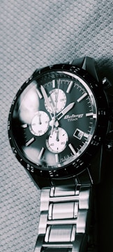 A silver chronograph wristwatch with a dark face and three subdials. It features a date window and has a metal link bracelet. The brand name on the face reads 'Octane Titan.' The watch is resting on a textured fabric surface.
