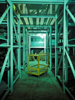 A dimly lit storage or warehouse area with metal shelving units lining both sides. A yellow metal cart with mesh sides is situated in the central aisle. Various items and boxes can be seen on the shelves and the floor in the background. The entire area has a greenish tint due to the lighting.