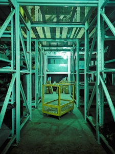 A dimly lit storage or warehouse area with metal shelving units lining both sides. A yellow metal cart with mesh sides is situated in the central aisle. Various items and boxes can be seen on the shelves and the floor in the background. The entire area has a greenish tint due to the lighting.