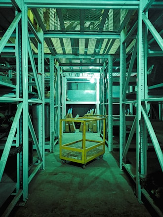 A dimly lit storage or warehouse area with metal shelving units lining both sides. A yellow metal cart with mesh sides is situated in the central aisle. Various items and boxes can be seen on the shelves and the floor in the background. The entire area has a greenish tint due to the lighting.