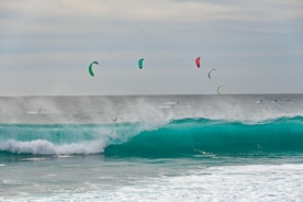 A group of kite surfers navigate large, teal waves with their kites soaring high in the sky. The ocean is rough, with mist and spray rising from the crashing waves. The kites are colorful, contrasting with the cloudy sky and the dark sea.