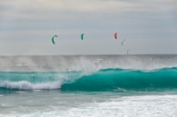 A group of kite surfers navigate large, teal waves with their kites soaring high in the sky. The ocean is rough, with mist and spray rising from the crashing waves. The kites are colorful, contrasting with the cloudy sky and the dark sea.