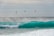 A group of kite surfers navigate large, teal waves with their kites soaring high in the sky. The ocean is rough, with mist and spray rising from the crashing waves. The kites are colorful, contrasting with the cloudy sky and the dark sea.
