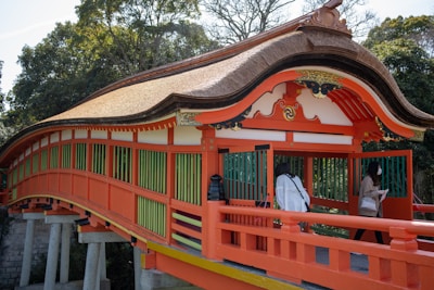 A vibrant, traditional bridge with a thatched roof is prominently displayed. The structure is painted in vivid orange and adorned with intricate designs. Green vertical railings contrast the orange frame. Trees with lush foliage can be seen in the background. Two people, wearing modern clothing and masks, are crossing the bridge.
