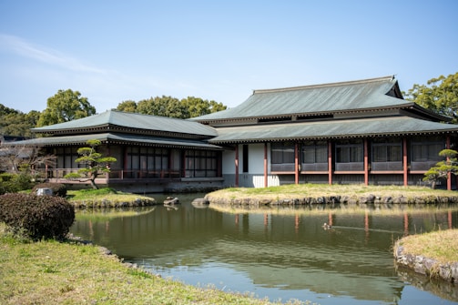 A traditional Japanese building with a green roof set beside a tranquil pond. The architecture features wooden elements and large windows, surrounded by manicured gardens and lush greenery.