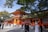An ornate, traditional Japanese shrine with vibrant red and orange hues is surrounded by lush green trees. A group of people, some dressed in traditional clothing, are gathered near the entrance, engaging in various activities. The shrine features intricate architectural details and a sloped, moss-covered roof.