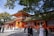 An ornate, traditional Japanese shrine with vibrant red and orange hues is surrounded by lush green trees. A group of people, some dressed in traditional clothing, are gathered near the entrance, engaging in various activities. The shrine features intricate architectural details and a sloped, moss-covered roof.