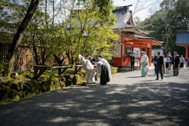 A group of people, some in traditional clothing, are gathered outside a Japanese temple on a sunny day. The temple features orange and white structures, and lush greenery surrounds the area. A few people appear engaged in a ritual or ceremony near a wooden structure.