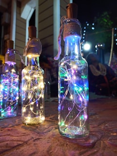 Glass bottles with cork lids are filled with string lights, creating a colorful and glowing effect. The lights emit a soft and vibrant glow in blue, green, and white hues. The setting appears to be outdoors with some twinkling lights in the background, adding a festive ambiance.