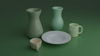 A collection of matte, pastel-colored ceramic kitchenware is arranged against a green backdrop. The items include two vases, a plate, a mug, and a cup. The vases are slightly different in shape and size, standing upright. The plate is flat and centrally positioned. The mug and cup are smaller and located at the front, displaying their handles.