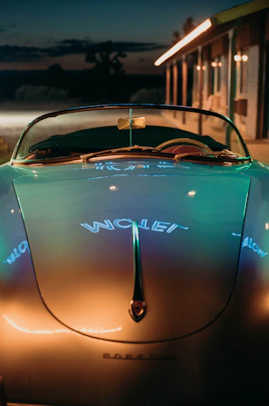 A shiny red 1980s convertible parked under glowing neon signs at dusk.