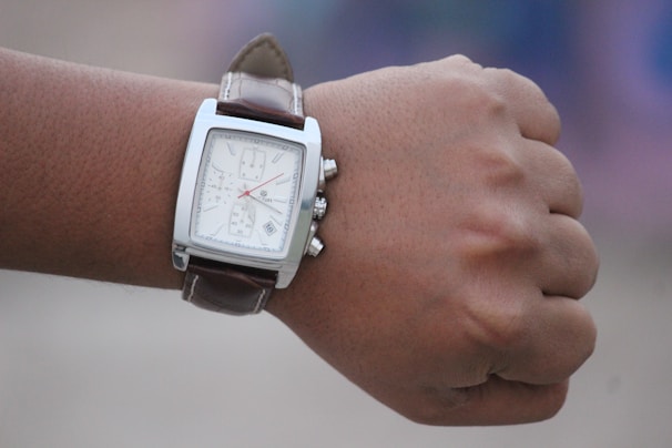 A close-up of a person's wrist wearing a square-faced wristwatch with a brown leather strap. The watch face is white with several dials and a date function.