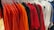 A collection of neatly arranged t-shirts on hangers, displaying a variety of colors including shades of orange, red, brown, white, and blue. The hangers are clear plastic, and the t-shirts are presented on a rack against a wooden floor.